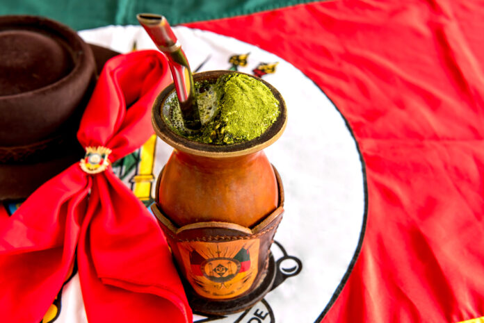 Chimarrão gaúcho em cuia tradicional com erva-mate, bomba de inox e fundo com bandeira do Rio Grande do Sul.