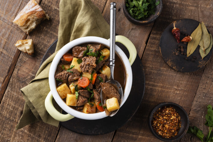 Puchero gaúcho servido em uma tigela com carne, mandioca e cenoura, acompanhado de temperos e pão rústico.