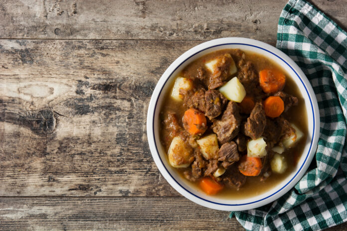 Prato de vaca atolada com carne, mandioca e cenoura em caldo, servido em tigela branca com fundo rústico.